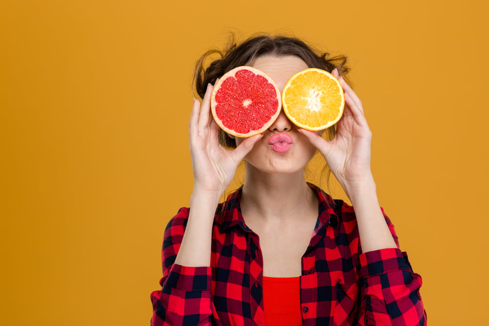 Funny playful young woman in checkered shirt holding halves of citrus fruits against her eyes and making duck face over yellow background Funny playful young woman in checkered shirt holding halves of citrus fruits against her eyes and making duck face over yellow background